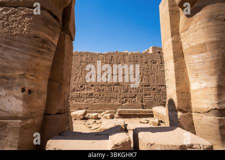 Antike ägyptische Hieroglyphen, die in eine Steinmauer im Tempel von Luxor in Ägypten gehauen wurden, eingerahmt von massiven Säulen unter einem hellblauen Himmel. Stockfoto