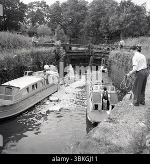 1950er Jahre, historisch, zwei schmale Boote in einer Kanalschleuse auf dem Land, oberes Tor wird geöffnet, Wasser tritt in die Schleuse ein, Mann auf dem Schleppweg neben der Schleuse, die Schleusenlande, mit Seil, England, Großbritannien. Stockfoto
