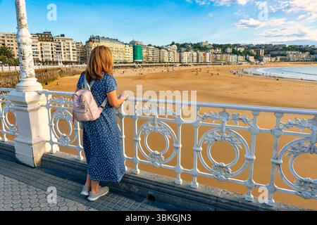 Weibliche Touristin auf dem Rücken, die den Blick auf den Strand La Concha bei Sonnenuntergang, San Sebastian, genießt. Stockfoto