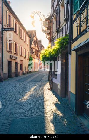 Die untergehende Sonne scheint durch ein Schild mit einer Weinbar und Fachwerkhäusern auf der Grand Rue im historischen Zentrum von Ribeauvillé, Elsass, Frankreich Stockfoto