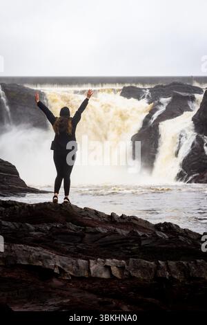 Eine Frau, die von hinten gesehen wird, steht vor den mächtigen Chaudière Falls und fängt einen Moment der Ehrfurcht in der Natur ein. Stockfoto