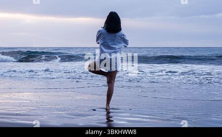 Das ruhige Bild zeigt eine Frau, die auf einem Bein in Yoga-Pose am Strand steht. Das dunkelblaue Meer und der bewölkte Himmel schaffen eine friedliche Atmosphäre. Stockfoto