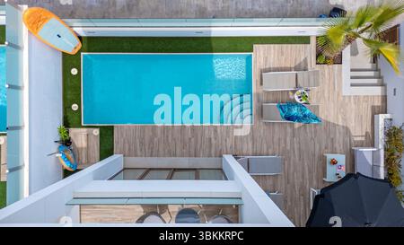 Blick von oben auf einen sauberen, türkisfarbenen Pool mit Grasrand, Holzterrasse und entspannenden Sonnenliegen in einer Villa. Stockfoto