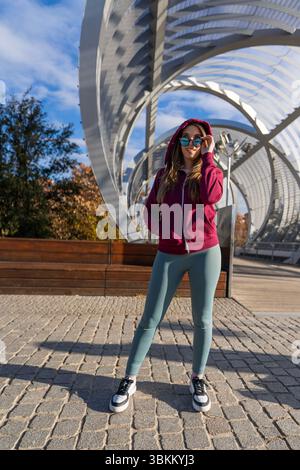 Junge Frau mit Sportbekleidung und Sonnenbrille, die auf einer modernen Fußgängerbrücke in madrid posiert Stockfoto