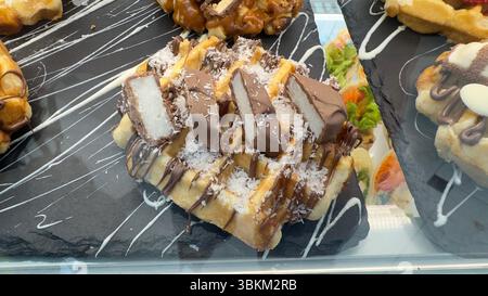 Nahaufnahme einer belgischen Waffel mit Schokolade, Kokosflocken und mit Kokosnuss gefüllten Schokoladentafeln auf einem schwarzen Schieferteller. Stockfoto