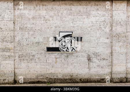 Das Wandgemälde aus der Serie "Réalité, Sortie de Secours" (Realität, Notausgang), entworfen von Marc-Antoine Mathieu, zeigt Comicfiguren, die aus der Wand zu platzen scheinen, Rue de Beaulieu, Angoulême, Nouvelle-Aquitaine, Frankreich. In dieser Tafel blickt ein Mann mit Hut und runden Brillen durch eine Kreuzschlitzlücke im gemalten Mauerwerk. In sechs Tafeln mit Sprechblasen erzählt die Serie eine vollständige Geschichte: "Siehst du etwas? Ja, schau. Es ist genau das, was ich befürchtet habe. Was, noch eine Unterwelt? Schlimmer als das. Es ist die Realität." Mathieu ist der Autor der Comicserie "Julius Corentin Stockfoto