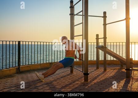 Fitter, gebräunter älterer, grauhaariger Mann, der bei Sonnenaufgang am Meer Trizepsdips an den Bars im Freien macht. Die Sonne reflektiert auf dem Wasser und wirft lange Schatten Stockfoto