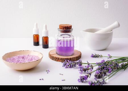 Glasflasche mit ätherischem Lavendelöl, Meersalz, Tropfflaschen, Mörtel und frischen Lavendelblüten auf weißem Hintergrund. Aromatherapiekonzept. Stockfoto
