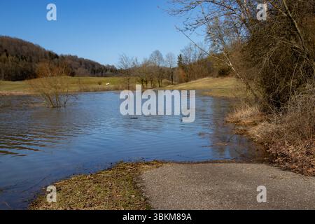 Eine Landstraße endet abrupt in einem überfluteten Gebiet, umgeben von einer ruhigen natürlichen Landschaft während eines Hochwasserereignisses, das durch saisonale Veränderungen verursacht wird. Stockfoto