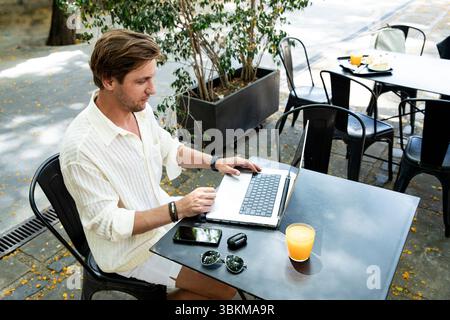 Freiberuflicher Mann, der in einem Open-Air-Café sitzt und einen Laptop benutzt. Entspannter und fröhlicher Mann, der fernab mit einem Drink auf dem Tisch arbeitet. Fernarbeiten im Innenraum Stockfoto
