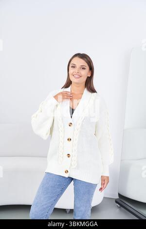 Lächelnde Frau in einem weißen Strickpullover mit großen Knöpfen und blauer Jeans in hellem Innenbereich, weißem Sofa teilweise sichtbar, modernes Design Stockfoto