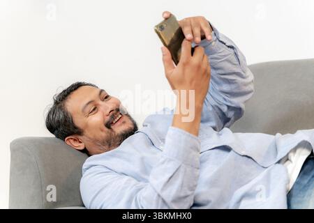 Junger asiatischer Mann, der bequem auf der Couch liegt und die Freizeit mit seinem Smartphone zu Hause genießt. Stockfoto