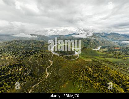 Aus der Vogelperspektive zieht sich ein sich windender Fluss durch üppig grüne und gelbe Herbstberge unter einem bewölkten Himmel Stockfoto