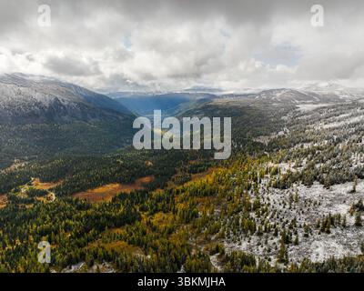 Eine spektakuläre Landschaft aus der Vogelperspektive zeigt, in der pulsierende Herbstwälder unter einem bewölkten Himmel in schneebedeckte Berge übergehen Stockfoto