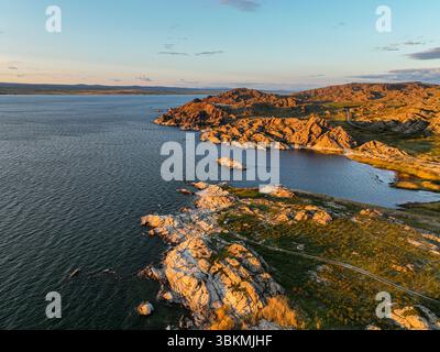 Ein Blick aus der Vogelperspektive auf eine zerklüftete, felsige Seeufer und Hügel, die vom sanften, warmen Licht der goldenen Stunde beleuchtet werden, mit Bergen in der Ferne Stockfoto