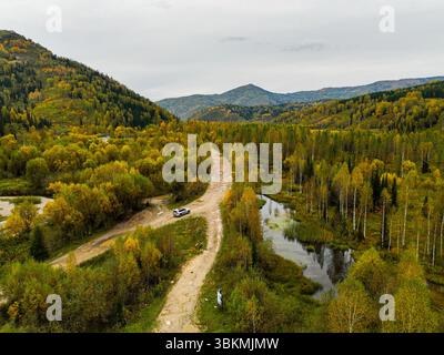 Ein Blick aus der Vogelperspektive fängt eine unbefestigte Straße mit einem Auto ein, die sich durch ein lebhaftes Herbsttal mit verschiedenen Bäumen und sanften Hügeln schlängelt Stockfoto