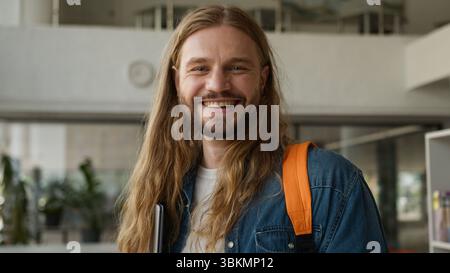 Porträt kaukasischer Studentenmann männlich positiv sorglos im Flur der Universitätsakademie, der Laptop hält, lächelnd, glücklich anschauend Stockfoto