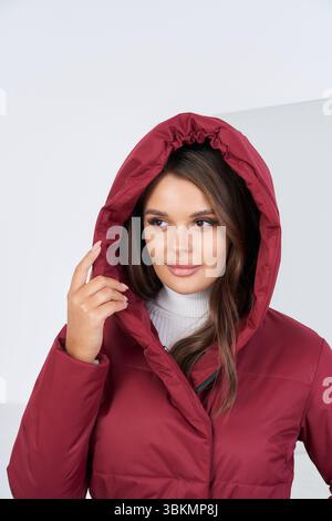 Frau in einer burgunderfarbenen Kapuzenjacke mit weißem Rollkragen darunter, seitlich guckend. Sie hat lange braune Haare und hält die Kapuze mit einer Hand Stockfoto