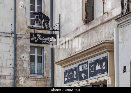 Ein Werbeschild für ein Fotostudio hängt in der Rue du Canton, Cognac, Nouvelle-Aquitaine, Frankreich. Das Schild für den Fotoladen "Les Photographes" zeigt die Silhouette eines Fotografen, der durch eine Kamera auf einem Stativ schaut. Darunter trägt eine schwarze Tafel den Namen in weißer Schrift, gehalten von einer schmiedeeisernen Halterung. Über dem Laden zeigt ein Schild das Wort "PHOTOS" und Piktogramme, die verschiedene Arten von Fotos darstellen, wie ein Porträt, eine Wolke, Berge, die Sonne und eine Blume. Das Haus trägt die Nummer 34. Stockfoto