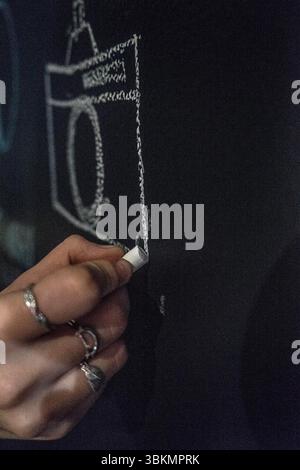 Eine Hand, die mit silbernen Ringen verziert ist, hält ein Stück weißer Kreide, die auf eine Tafel gezeichnet ist. Die Szene fängt einen Moment der Kreativität und Kunstfertigkeit ein. Stockfoto