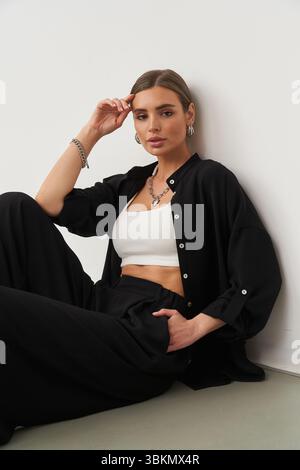 Frau saß entspannt auf dem Boden, trug ein schwarzes offenes Hemd über einem weißen, abgeschnittenen Oberteil, schwarze Hosen und Silberschmuck. Ihre Hand liegt auf ihr Stockfoto