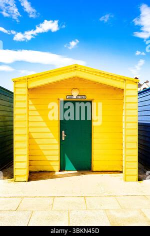 Hellgelbe Strandhütte mit grüner Tür unter einem leuchtend blauen Himmel mit weißen Wolken, umgeben von anderen farbenfrohen Hütten Stockfoto