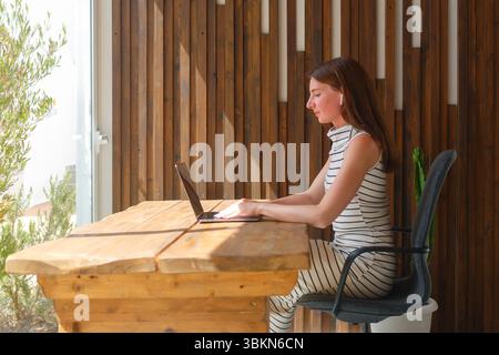 Eine Frau sitzt an einem hölzernen Schreibtisch, tippt auf einem Laptop in einem hellen, luftigen Raum, umgeben von Holzwänden und natürlichem Licht. Stockfoto
