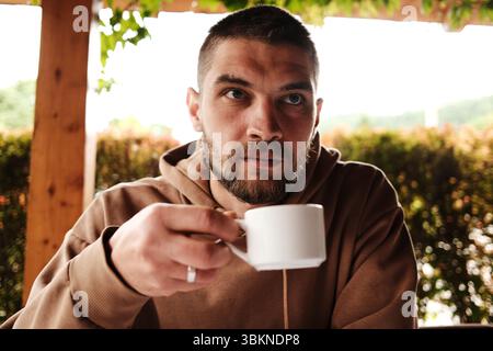Bärtiger Mann in einem braunen Hoodie, der Kaffee in einem traditionellen serbischen Restaurant trinkt. Tageslicht und gemütliche Holzumgebung Stockfoto