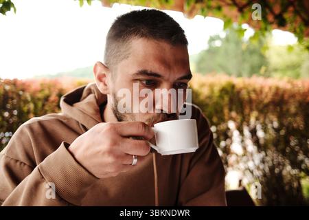 Bärtiger Mann in einem braunen Hoodie, der Kaffee in einem traditionellen serbischen Restaurant trinkt. Tageslicht und gemütliche Holzumgebung. Stockfoto