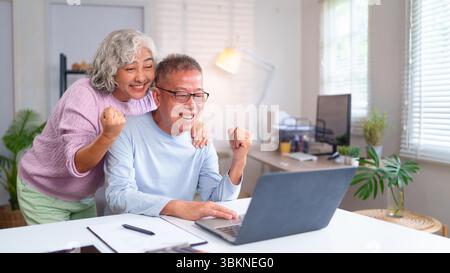 Ein älteres asiatisches Paar drückt Freude in seinem Heimbüro aus, während es einen Laptop benutzt. Dies fängt ihren glücklichen Ruhestand, ihren Erfolg und ihren fröhlichen Moment ein Stockfoto