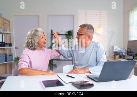Fröhliches, älteres asiatisches Paar feiert Erfolg mit einer High Five im Home Office. Glückliche Senioren genießen gemeinsam ihren Lebensstil im Ruhestand Stockfoto