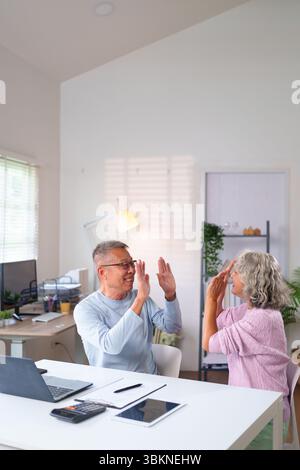 Glückliches älteres asiatisches Paar feiert einen Sieg mit einer hohen fünf im Home Office und zeigt Freude. Sie planen ihren Ruhestand mit technischen Geräten. Stockfoto