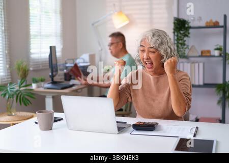 Eine ältere asiatische Frau freut sich über den Erfolg ihres Laptops im Heimbüro, während ein älterer Mann hinter ihr arbeitet. Erlebe ihren Sieg Stockfoto