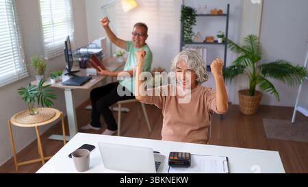 Ein überfröhliches älteres asiatisches Paar feiert Erfolge in ihrem Home Office, drückt Glück aus und erreicht gemeinsam Ziele. Ein fröhliches Rentenkonzept Stockfoto
