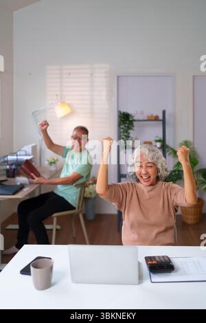 Ein glückliches älteres asiatisches Paar, das zu Hause mit Laptop und Dokumenten einen Erfolg feiert. Sie erreichen gemeinsam Ziele, während sie ihre Altersvorsorgefonds verwalten. Stockfoto
