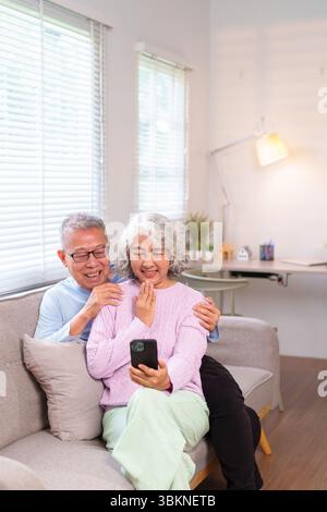 Asiatisches älteres Paar sitzt zusammen auf Sofa mit Smartphone für Videoanrufe lächelnd zu Hause glücklicher älterer Mann und Frau, die mit dem Mobiltelefon Spaß haben Stockfoto