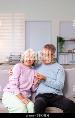 Dieses Bild zeigt asiatische ältere Ehepaare, die zu Hause gute Zeit miteinander verbringen. Sie entspannen auf einem Sofa, halten Hände und lächeln vor Liebe und Happy Stockfoto