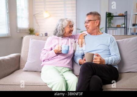 Ein herzerwärmendes älteres asiatisches Paar sitzt bequem auf der Couch, hält Kaffeetassen, zeigt Zuneigung und genießt ihre goldenen Jahre in ihrem Ko Stockfoto