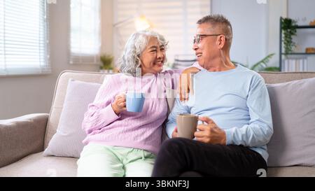 Ein älteres asiatisches Paar teilt sich einen liebevollen Moment, während es zu Hause Kaffee auf dem Sofa genießt. Sie lächeln und lachen und zeigen die Freuden des Ruhestands und c Stockfoto