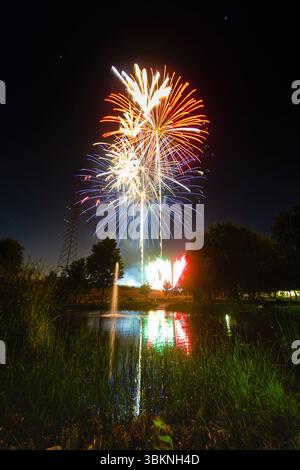 Helles Feuerwerk in verschiedenen Farben über einem ruhigen Teich, das sich nachts lebhaft im Wasser spiegelt. Stockfoto