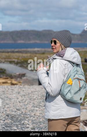 Frau in weißer Jacke und grauer Mütze, die einen hellblauen Rucksack mit gelbem Regenschutz hält und auf einem felsigen Pfad im Naturschutzgebiet Teriberka steht Stockfoto