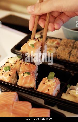Nahaufnahme von Sushi-Lachsrollen in einem schwarzen Tablett zum Mitnehmen mit einer Hand, die Stäbchen hält. Zu den Belägen gehören Sesamsamen und Gemüse. Perfekt für Food-Blogs Stockfoto