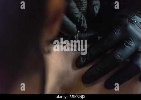 Ein Tätowierer mit schwarzen Handschuhen arbeitet an einem Tattoo auf dem Rücken einer Person. Der Künstler fügt dem Design sorgfältig Details hinzu. Stockfoto