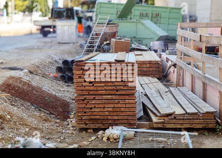 Eine Szene einer Baustelle mit mehreren Stapeln von Holzdielen im Vordergrund, Leitern, Rohren und anderen Baumaterialien und schweren Fahrzeugen Stockfoto
