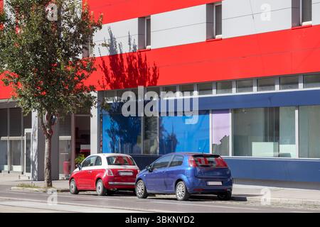 Zwei Autos – ein roter mit weißem Dach und ein blauer – parken auf der Straße neben einem modernen Gebäude mit roten, grauen und blauen Details. Stockfoto