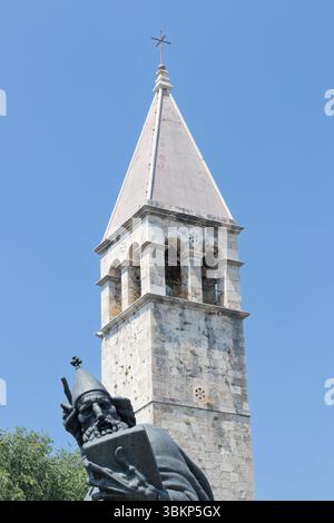SPLIT, KROATIEN, 08.06.2018: Eine ruhige Szene mit einem historischen Glockenturm neben einer hohen Statue, umgeben von üppigem Grün, unter einem klaren blauen Himmel, Stockfoto