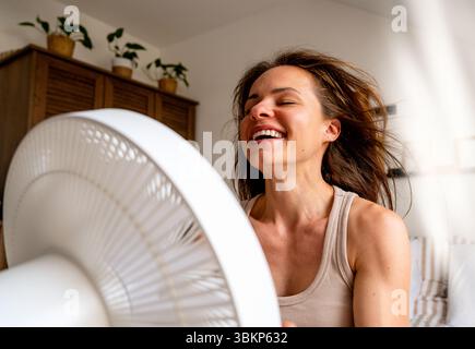 Glückliche junge Frau lächelt und genießt einen erfrischenden Luftstrom vom elektrischen Ventilator an heißen Sommertagen Stockfoto