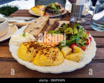 Gourmet-Frühstücksteller mit benedict-Eiern, Lachs, frischem Salat, Käse und Sauerteigbrot, serviert auf einer Café-Terrasse. Ein gesundes und köstliches Stockfoto