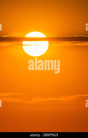 Goldene Sonne, die bei Sonnenauf- oder -Untergang durch dünne Wolken am leuchtenden orangefarbenen Himmel leuchtet und eine warme, traumhafte Atmosphäre schafft Stockfoto