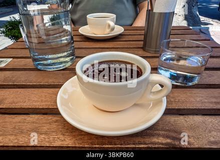 Morgenkaffee auf einer Sonnenterrasse im Freien. Nahaufnahme von Americano-Kaffee und Wasser auf einem Holztisch. Gemächlicher Nachmittag in einem Café: Zwei Tassen Cof Stockfoto